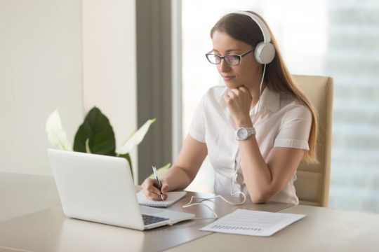 Woman wearing headphones and taking notes while looking at a laptop.