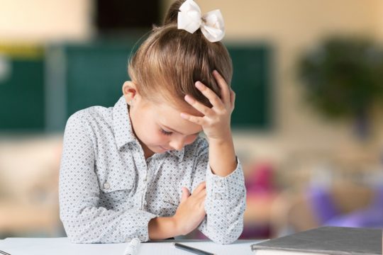 Young girl holding her head while sitting at a desk with a notebook and pencil.