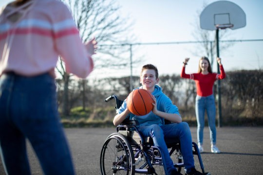 Young boy in a wheelchair playing basketball with two girls.