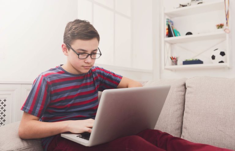 Young boy with glasses sitting on a couch working on a laptop.