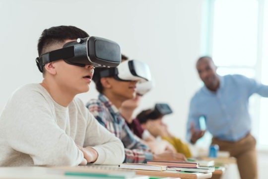 Group of students sitting at tables wearing virtual reality goggles.