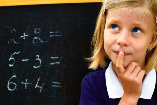 Young girl standing in front of a chalkboard with math problems thinking.