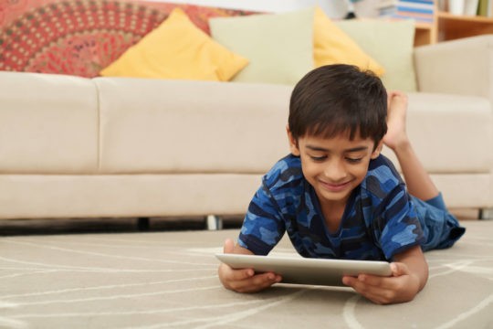 Young boy lying on the ground smiling and using a tablet.