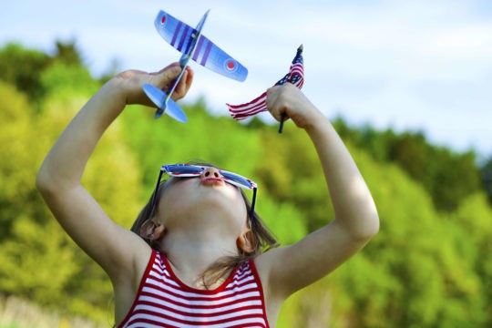 A little girl playing outside with a toy airplane and a small American flag.
