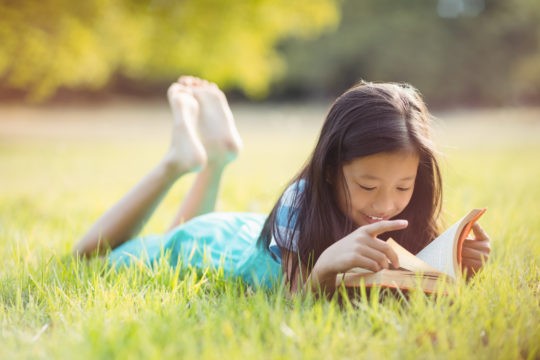 Young girl lying in the grass reading a book.