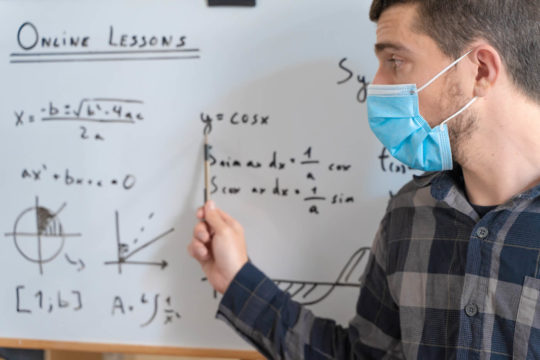 A teacher wearing mask and pointing to a whiteboard on video.