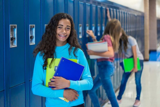 Teenage girl smiling and holding notebooks in a school hallway.