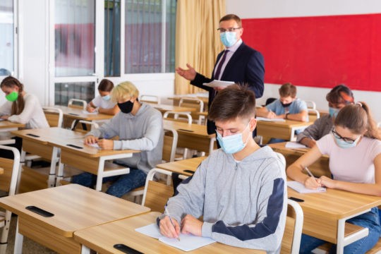 Teacher walking around a classroom while students work at their desks, all wearing masks.