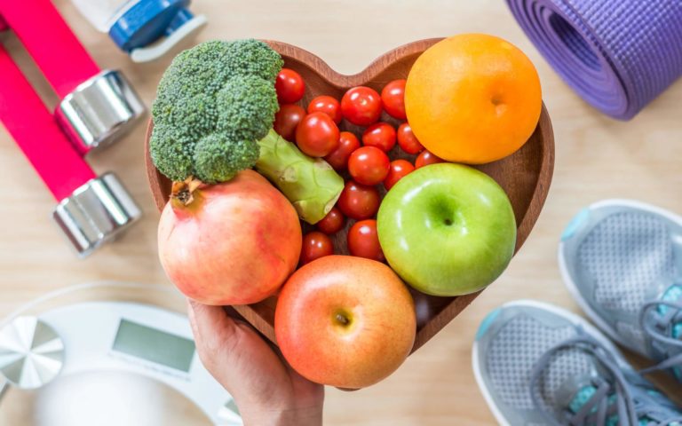 Heart-shaped bowl of colorful fruits and vegetables surrounded by fitness items.