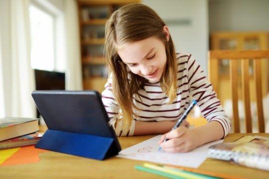 Young girl using a tablet and drawing at a table.