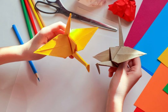 Children’s hands make an origami crane, above a white background with various school supplies.