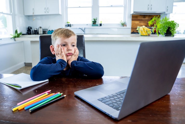Young boy sitting at a kitchen table with a laptop looking very bored.
