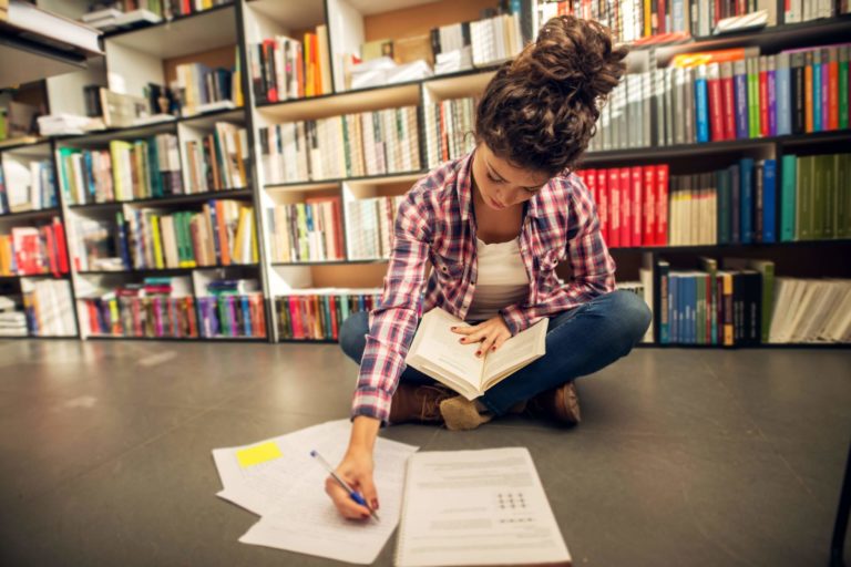 Female student sitting in a library reading a book and taking notes.