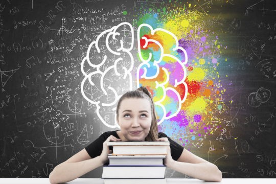 A girl sits resting her head on a stack of books while looking up at a brain sketch on the blackboard behind her.