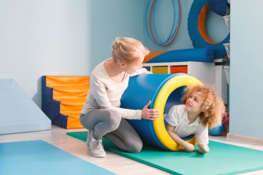 Young boy crawling through a tunnel in a sensory room with a teacher monitoring.