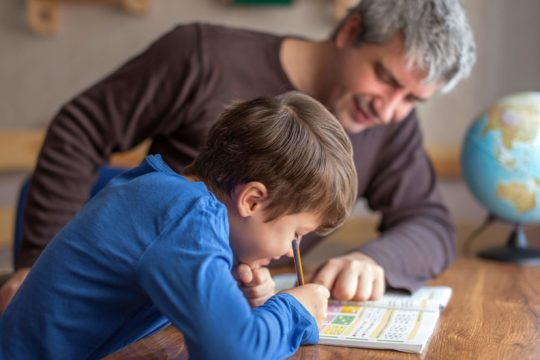 Father sitting with his son at a table working on a school workbook.