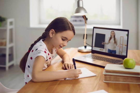 Young girl writing at a table while on video chat with her teacher