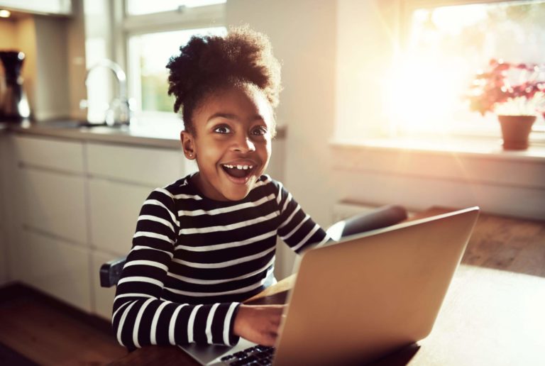 Young girl sitting at a table working on a laptop looking very excited.