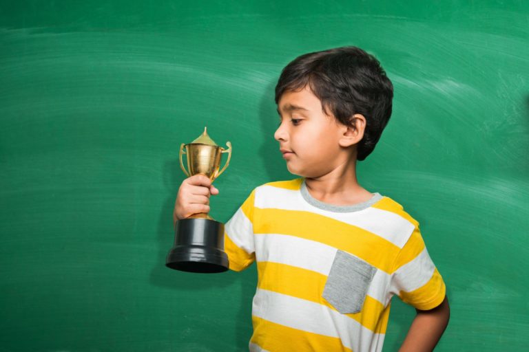 Young boy holding a trophy standing in front of a chalkboard.