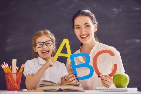Young girl holding up letters of the alphabet with teacher in front of chalkboard