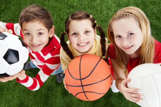Young kids looking up and smiling while holding different sports balls