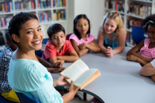 Female teacher reading a book to a group of students at a table